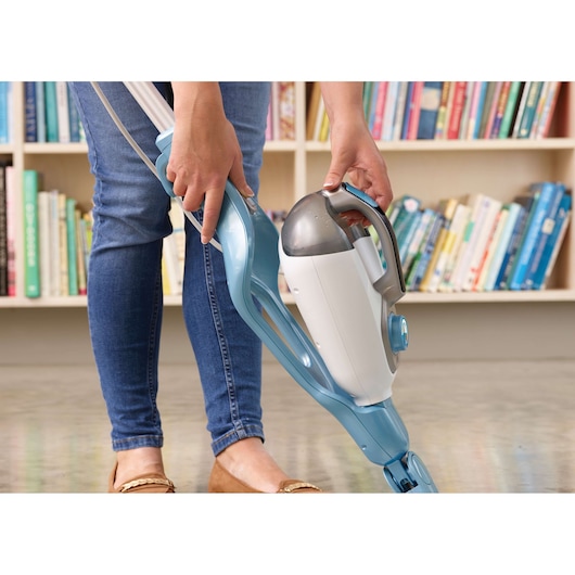 A person is using a BLACK+DECKER steam mop with a detachable handheld unit on a hard floor, in front of a bookshelf filled with books.