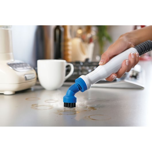 Close-up of a hand using a BLACK+DECKER steam cleaning tool with a blue brush attachment to clean stains on a kitchen countertop. A white mug and kitchen appliances are visible in the background.