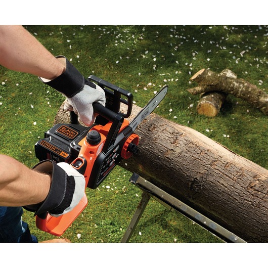 A person wearing gloves operates a BLACK+DECKER cordless chainsaw to cut a log on a metal stand outdoors, with additional logs and green grass in the background.