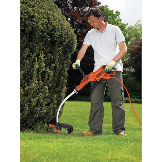 A person using a BLACK+DECKER electric grass trimmer (model GL7033) to trim grass along the edge of a hedge in a garden.