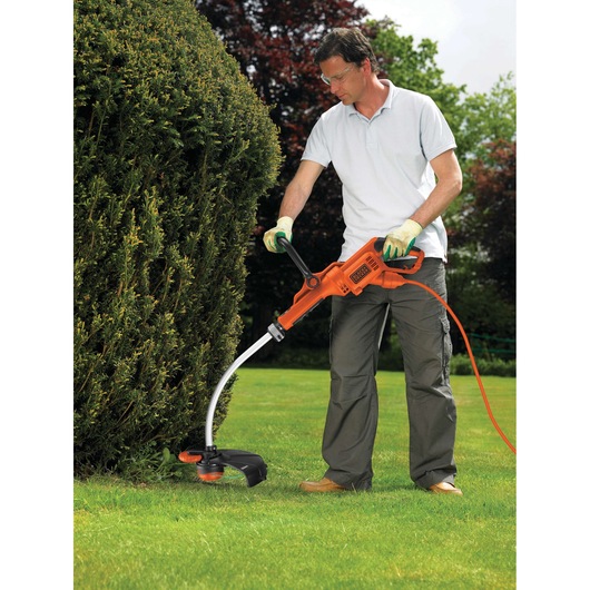 A person using a BLACK+DECKER GL9035 electric grass trimmer to cut grass near a bush in a garden. The individual is wearing gloves and casual outdoor clothing.