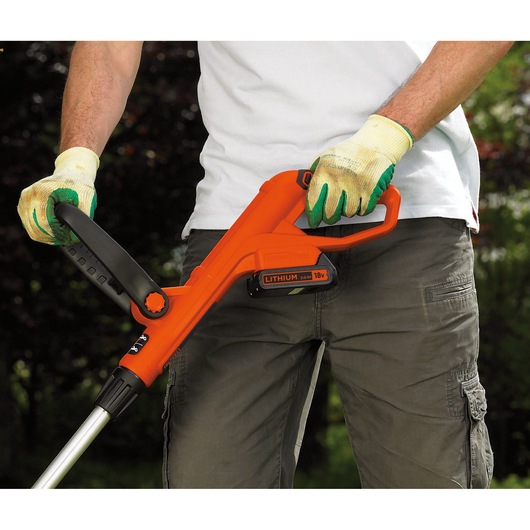 A person wearing gardening gloves operates an orange BLACK+DECKER cordless grass trimmer powered by an 18V lithium battery, outdoors.