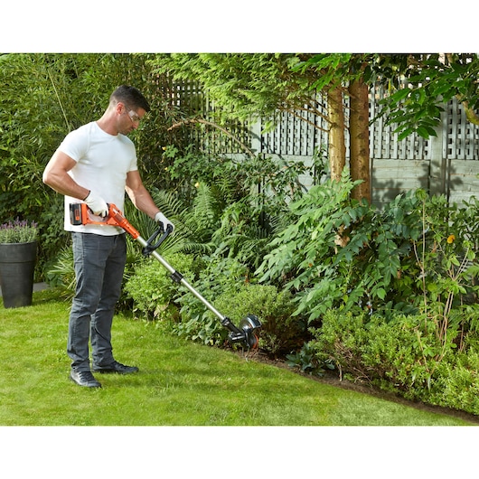 A person using a BLACK+DECKER cordless grass trimmer in a garden, trimming the grass along the edge of a flower bed. The person is wearing gloves and casual clothing.