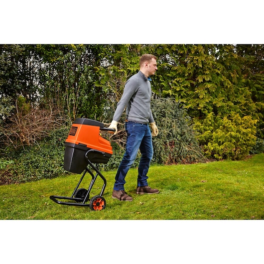 A person wearing gloves is pulling a BLACK+DECKER GS2400 garden shredder with wheels across a grassy lawn. The shredder is orange and black in color, and there are trees and shrubs in the background.