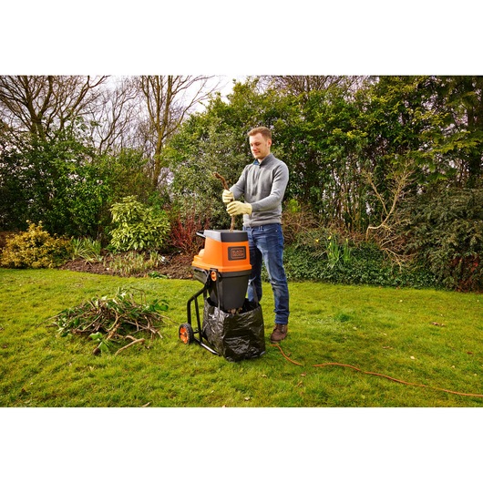 A person wearing gloves uses a BLACK+DECKER GS2400 garden shredder outdoors. The machine is shredding branches and twigs into a black collection bag. The person stands on a grassy lawn with a pile of garden debris nearby, surrounded by bushes and trees.