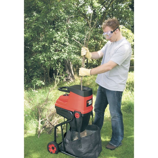 A person wearing gloves uses a BLACK+DECKER GS2400 garden shredder to shred branches outdoors. The machine is red and black, with branches being fed into the top and shredded material collected in a black bag below.