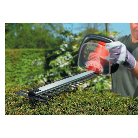 A person using a BLACK+DECKER electric hedge trimmer to trim a hedge outdoors, wearing protective gloves.