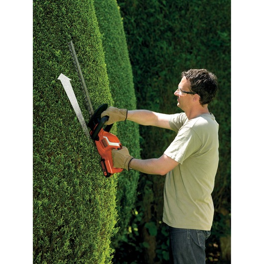 A person wearing gloves using a BLACK+DECKER cordless hedge trimmer to trim a tall, dense hedge outdoors. The hedge trimmer is orange and black, and an arrow indicates the direction of cutting.