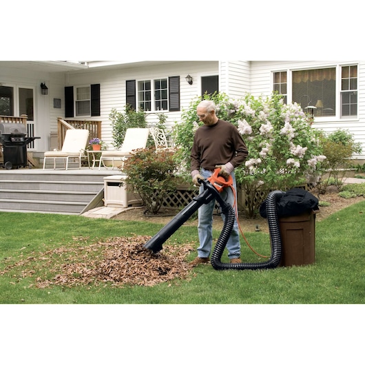 Person using BLACK+DECKER GW3050 leaf blower vacuum on lawn with brown garden bin and black hose attachment.