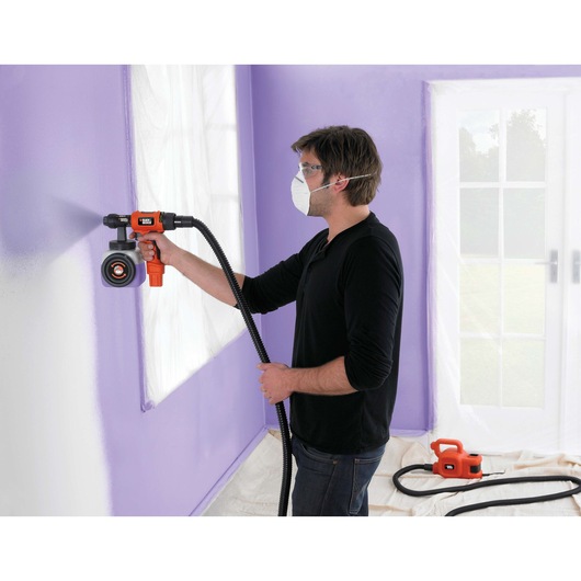 A person using a BLACK+DECKER HVLP400 paint sprayer to apply purple paint to an indoor wall. The individual is wearing a mask, and nearby windows and floor are protected with plastic sheeting. The paint sprayer's hose is connected to a base unit on the floor.
