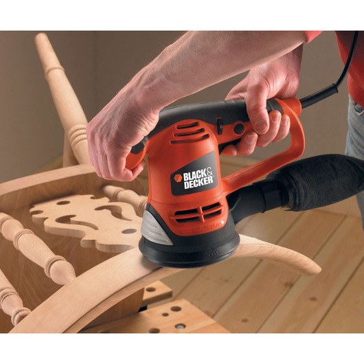 A person uses a BLACK+DECKER electric sander to smooth a wooden chair component on a workbench.