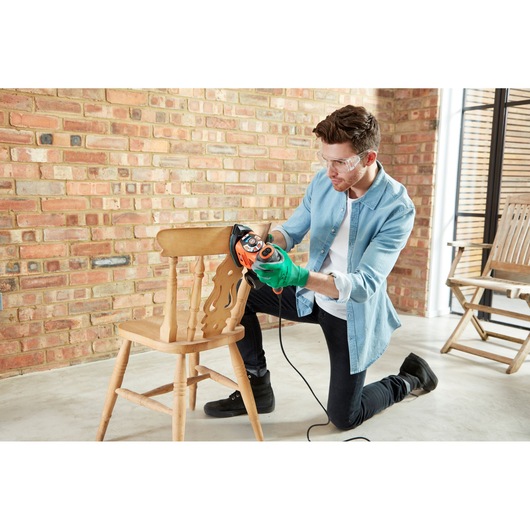 A person wearing green gloves is kneeling and using a BLACK+DECKER KA280LSA electric sander to sand a wooden chair in front of a brick wall.
