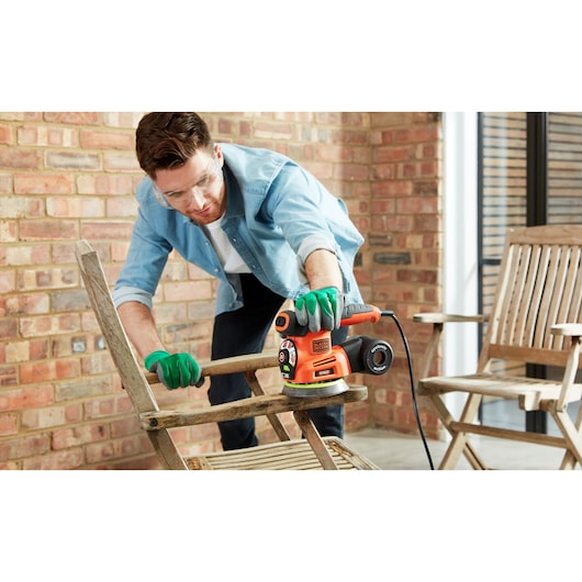 A person wearing green gloves is using a BLACK+DECKER KA280 sander to refurbish a wooden chair on a patio, with brick walls and additional wooden chairs in the background.