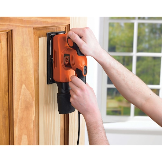 A person is using a BLACK+DECKER electric sander to sand a wooden door, with a window visible in the background.