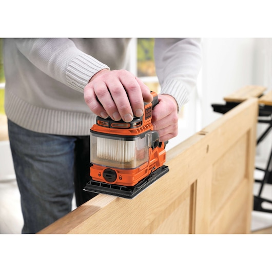 A person is using a BLACK+DECKER KA330E orange electric sander to sand the edge of a wooden door. The tool has a dust collection compartment and is held firmly during the sanding process.