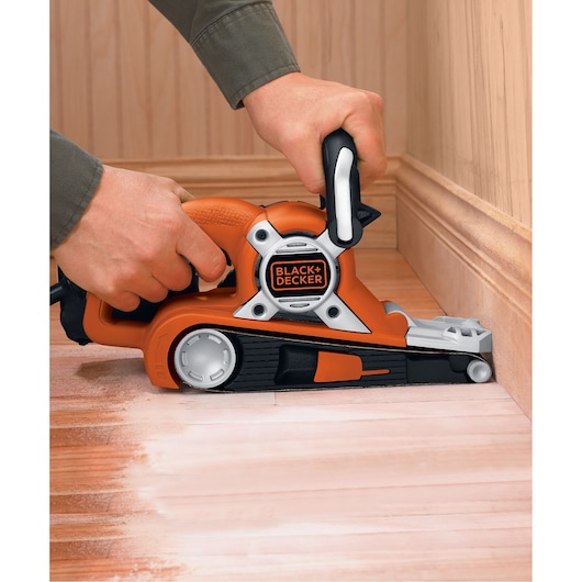 A person using a BLACK+DECKER KA88 belt sander to sand a wooden floor near the edge of a wall.