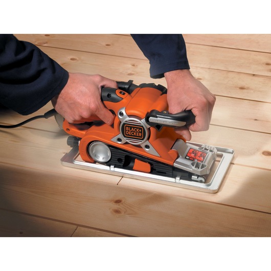 A person using a BLACK+DECKER electric belt sander to smooth wooden floorboards.