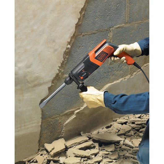 A person wearing gloves is using a BLACK+DECKER electric hammer drill with a chisel attachment to remove plaster from a wall, with broken debris on the floor.