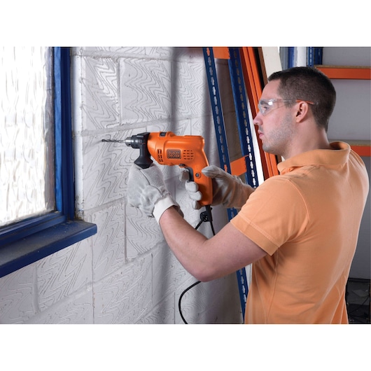 A person wearing protective gloves and an orange shirt is using a BLACK+DECKER electric drill (model KR504RE) to drill into a white brick wall near a window.