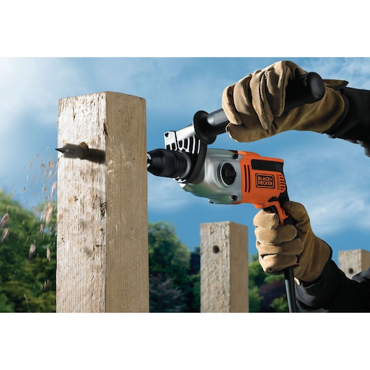 A person wearing protective gloves uses a BLACK+DECKER electric drill to bore a hole into a wooden post outdoors, with wood shavings visible.
