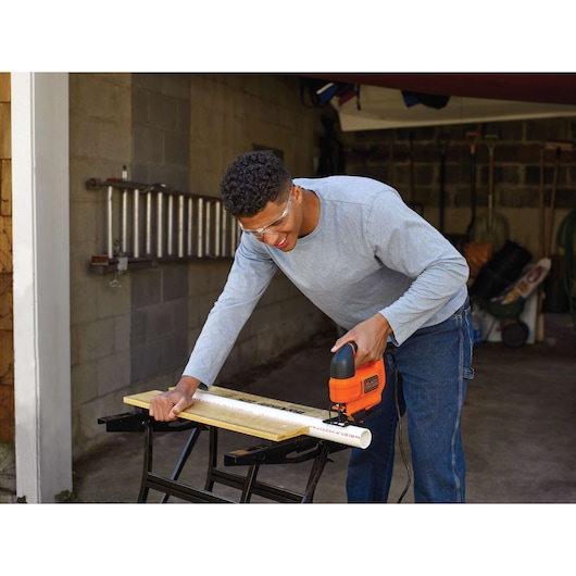 A person using a BLACK+DECKER jigsaw (KS701E) to cut a white PVC pipe on a workbench in a garage. The person's face is blurred for privacy.