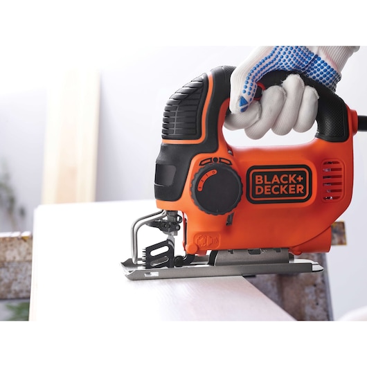 A close-up view of a BLACK+DECKER electric jigsaw being operated with a gloved hand, cutting through a piece of wood.