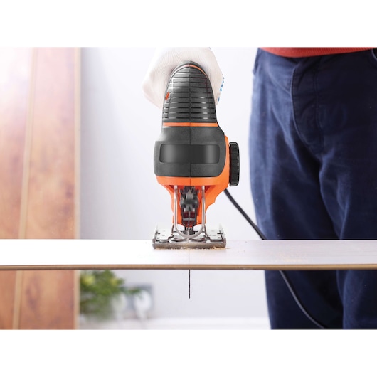 A person using a BLACK+DECKER electric jigsaw to cut through a wooden board, with a close-up view of the tool and the wood.