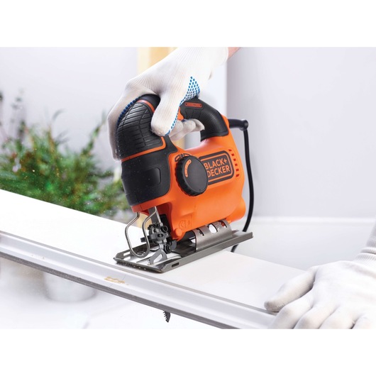 A person wearing white gloves uses a BLACK+DECKER orange and black electric jigsaw to cut a white piece of material.