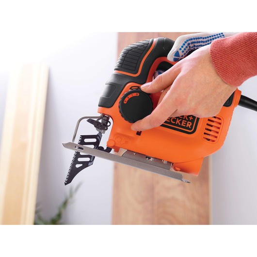 Close-up of a person adjusting the settings on an orange BLACK+DECKER jigsaw, with the saw blade visible and wooden boards in the background.