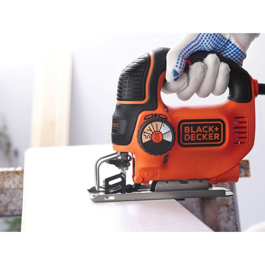A close-up of a BLACK+DECKER electric jigsaw being used by a person wearing white and blue safety gloves to cut a piece of wood.