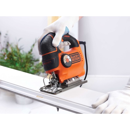 A person wearing white gloves uses an orange and black BLACK+DECKER jigsaw to cut a piece of wood indoors. The jigsaw is being guided along a straight edge.