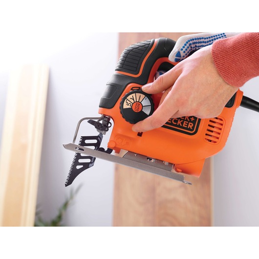 Close-up of a person adjusting the settings on an orange and black BLACK+DECKER jigsaw tool, with visible blade and control knob.