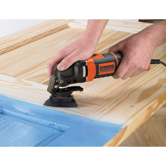 A BLACK+DECKER MT300KA multi-tool is being used to sand the surface of a wooden door with blue paint.