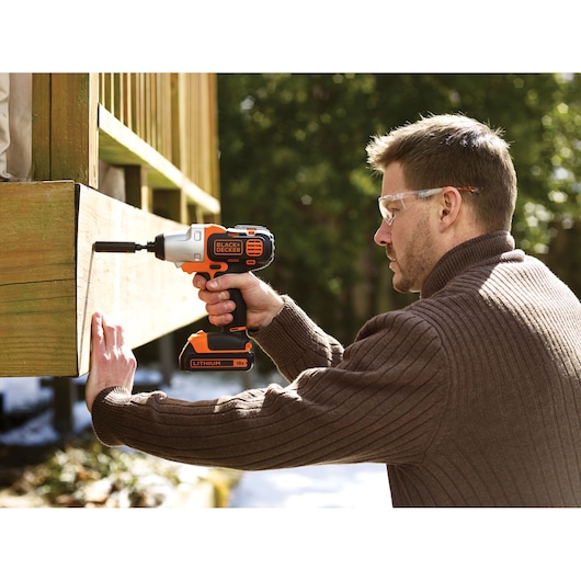 A person using a BLACK+DECKER 18V Lithium cordless power drill to drive a screw into a wooden structure outdoors. The person's face is blurred for privacy.