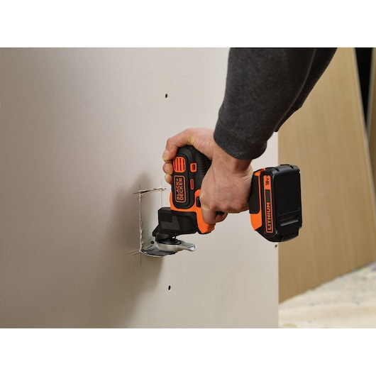 A person uses a BLACK+DECKER MTOS4 multi-tool with a battery pack to cut a square opening in a drywall panel.