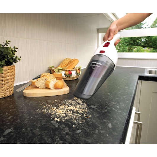 A handheld BLACK+DECKER vacuum cleaner is being used to clean breadcrumbs from a kitchen countertop. There is a wooden cutting board with sliced bread and a basket of baguettes in the background.