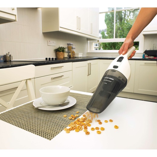 A BLACK+DECKER handheld vacuum cleaner is being used to pick up spilled cereal from a white kitchen table, next to a bowl and spoon in a modern kitchen setting.