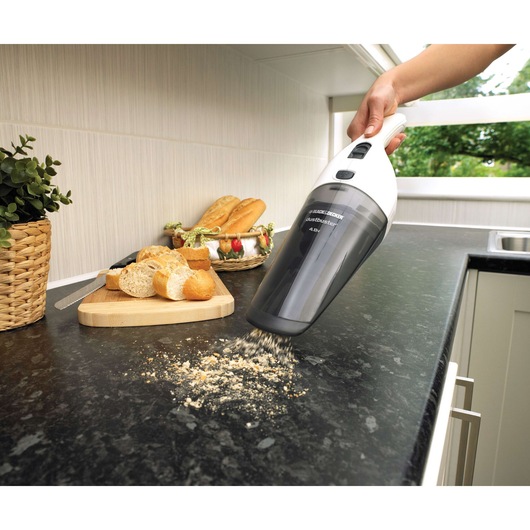 A handheld BLACK+DECKER dustbuster vacuum is being used to clean bread crumbs from a kitchen countertop. Nearby, there is a chopping board with sliced bread, a basket of bread, and a potted plant.