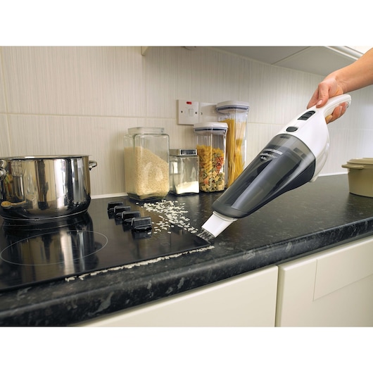 A BLACK+DECKER handheld vacuum cleaner is being used to clean spilled rice from a kitchen countertop and stove. There are jars containing rice, pasta, and other ingredients in the background.