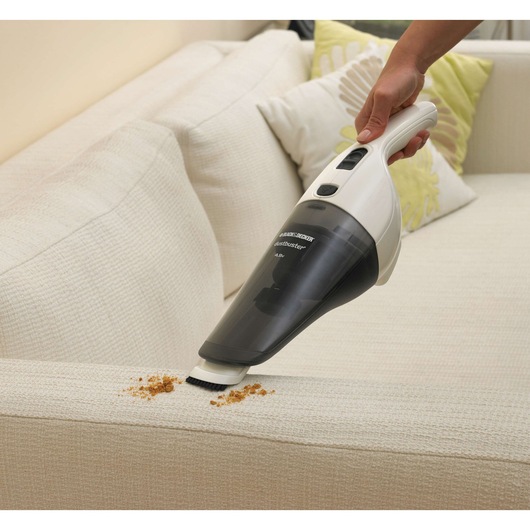A person uses a BLACK+DECKER handheld vacuum cleaner (model NV4820N) to clean crumbs from the surface of a light-colored sofa.