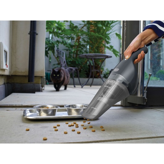A person uses a BLACK+DECKER handheld vacuum cleaner to pick up scattered pet food near metal bowls, with a black cat in the background on a patio.