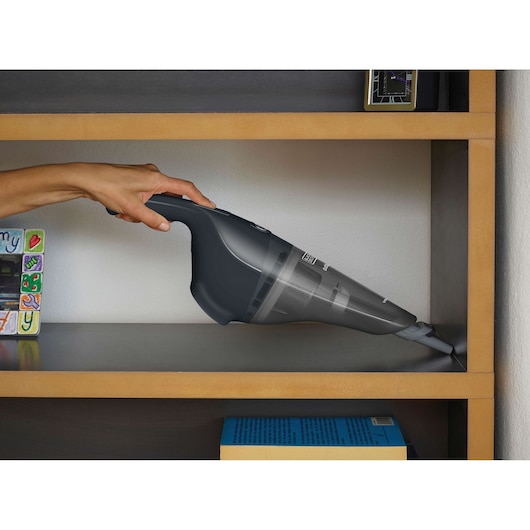 A person uses a BLACK+DECKER handheld vacuum cleaner to clean the corner of a wooden shelf.