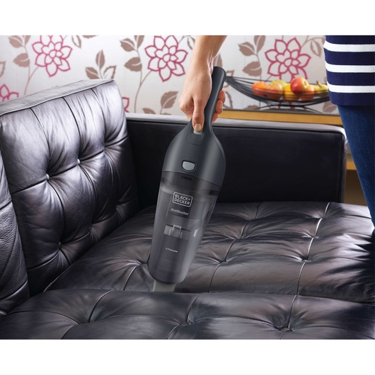 A person is using a BLACK+DECKER handheld vacuum cleaner (model NVB215WA) to clean a black leather sofa in a living room. There is a bowl of fruit in the background on a table and floral wallpaper on the wall.