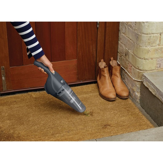 A person using a BLACK+DECKER handheld vacuum cleaner (model NVB215WA) to clean a brown doormat at the entrance of a wooden door, with a pair of tan boots placed beside the door.