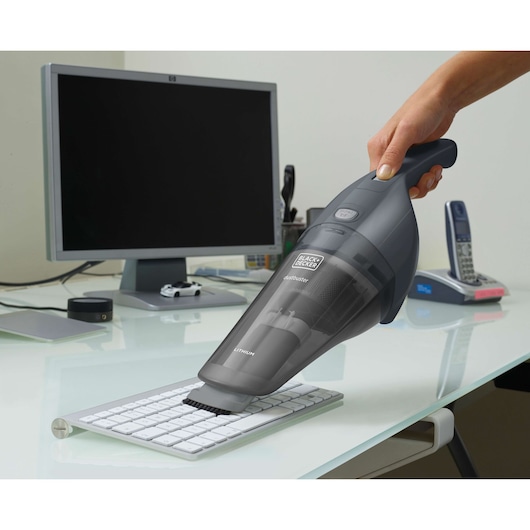A BLACK+DECKER handheld vacuum cleaner is used to clean a computer keyboard on a modern office desk. A monitor, telephone, and other office items are visible in the background.