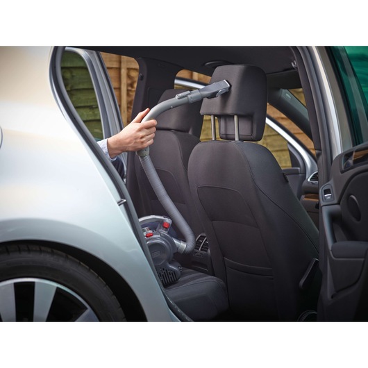 A person is using a BLACK+DECKER portable vacuum cleaner to clean the interior of a car, specifically the headrest area of the front seat. The vacuum is equipped with a flexible hose and nozzle, suitable for reaching tight spaces inside vehicles.
