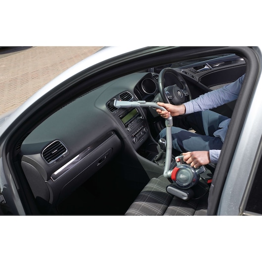 A person is using a BLACK+DECKER portable vacuum cleaner (model PD1200AV) to clean the interior of a car. The vacuum cleaner is handheld and features a flexible hose, making it ideal for reaching tight spaces inside the vehicle.