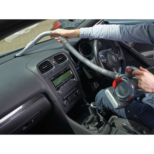 A person uses a BLACK+DECKER portable vacuum cleaner (model PD1200AV) to clean the dashboard area inside a car. The vacuum has a flexible hose and is powered by a plug-in cable.