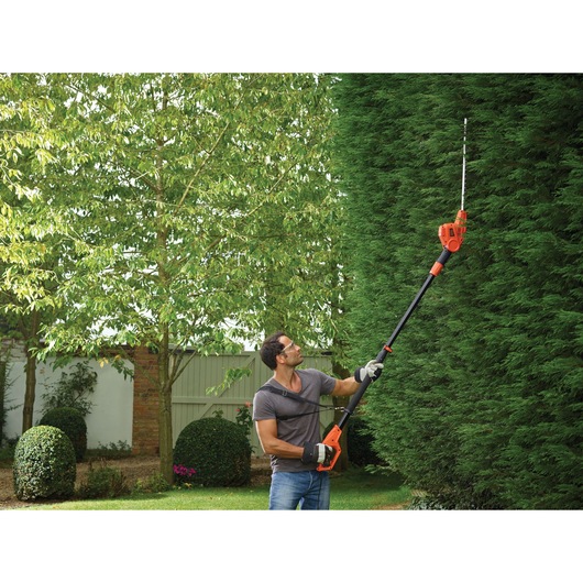 A person uses a BLACK+DECKER pole hedge trimmer (model PH5551) to trim tall hedges in a garden. The tool has an extended pole for reaching high branches, and the person is wearing gloves and a harness for safety.