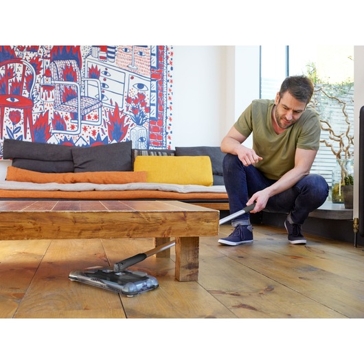 A person using a BLACK+DECKER cordless vacuum cleaner (model PSA215B) to clean under a wooden coffee table in a modern living room with colorful cushions and an abstract wall hanging.